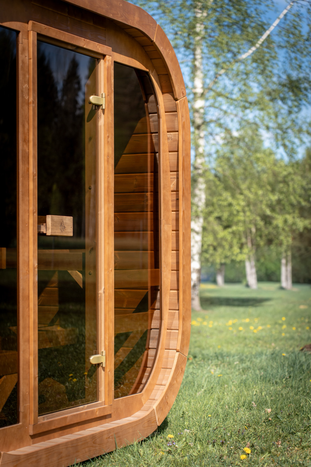 Close view of the door edge on a PlusLife Max Traditional Sauna 2.0: two brass hinges on a timber-framed glass door, horizontal timber staves curving around the rounded corner of the cabin, and a timber bench visible through the glass